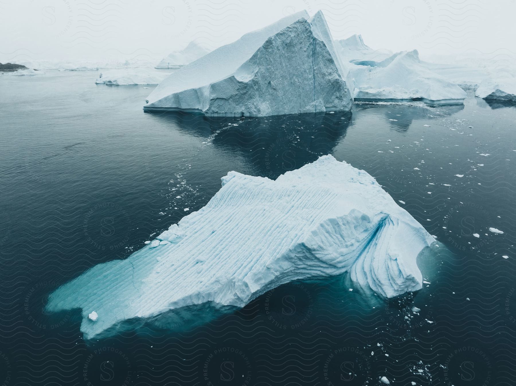 Icebergs and pieces of ice float in arctic water under a white hazy sky