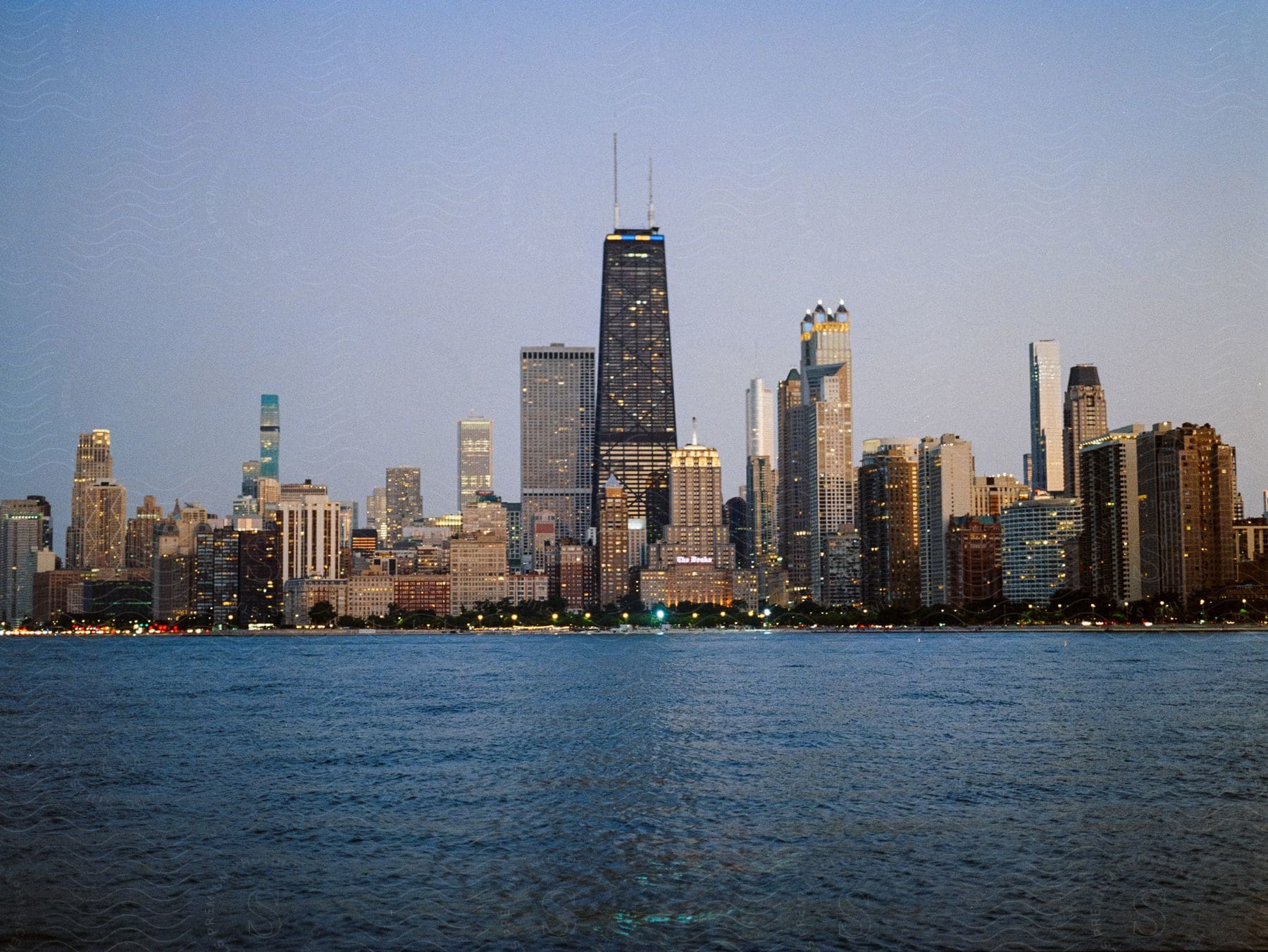 Various sized city buildings on a waterfront with the buildings lit up at dusk