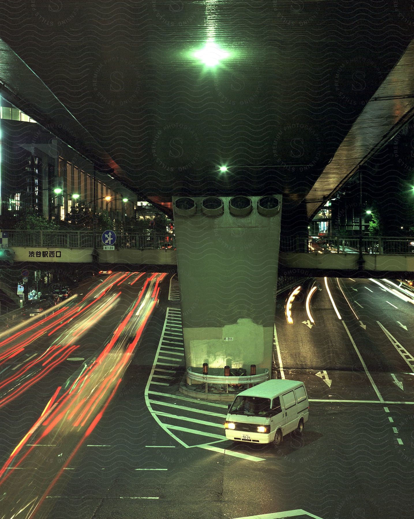 A van waits to turn under an overpass at night in a metropolis with streaks of blurred light from moving traffic