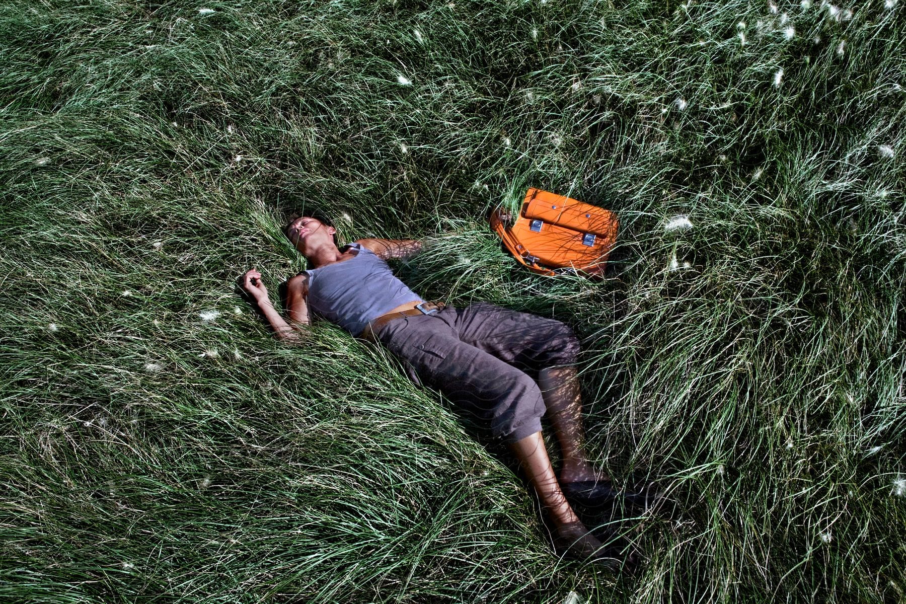 A man reclining on the grass in a field accompanied by an orange bag resting beside him