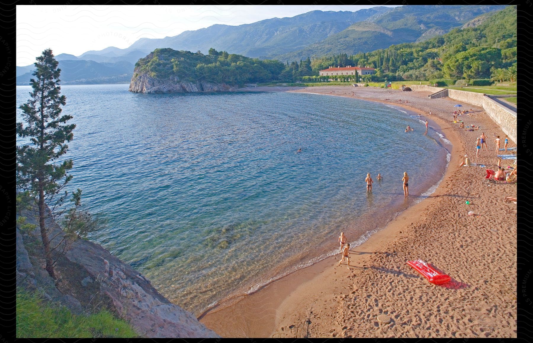 People on beach and in water with building near promontory and mountains in distance