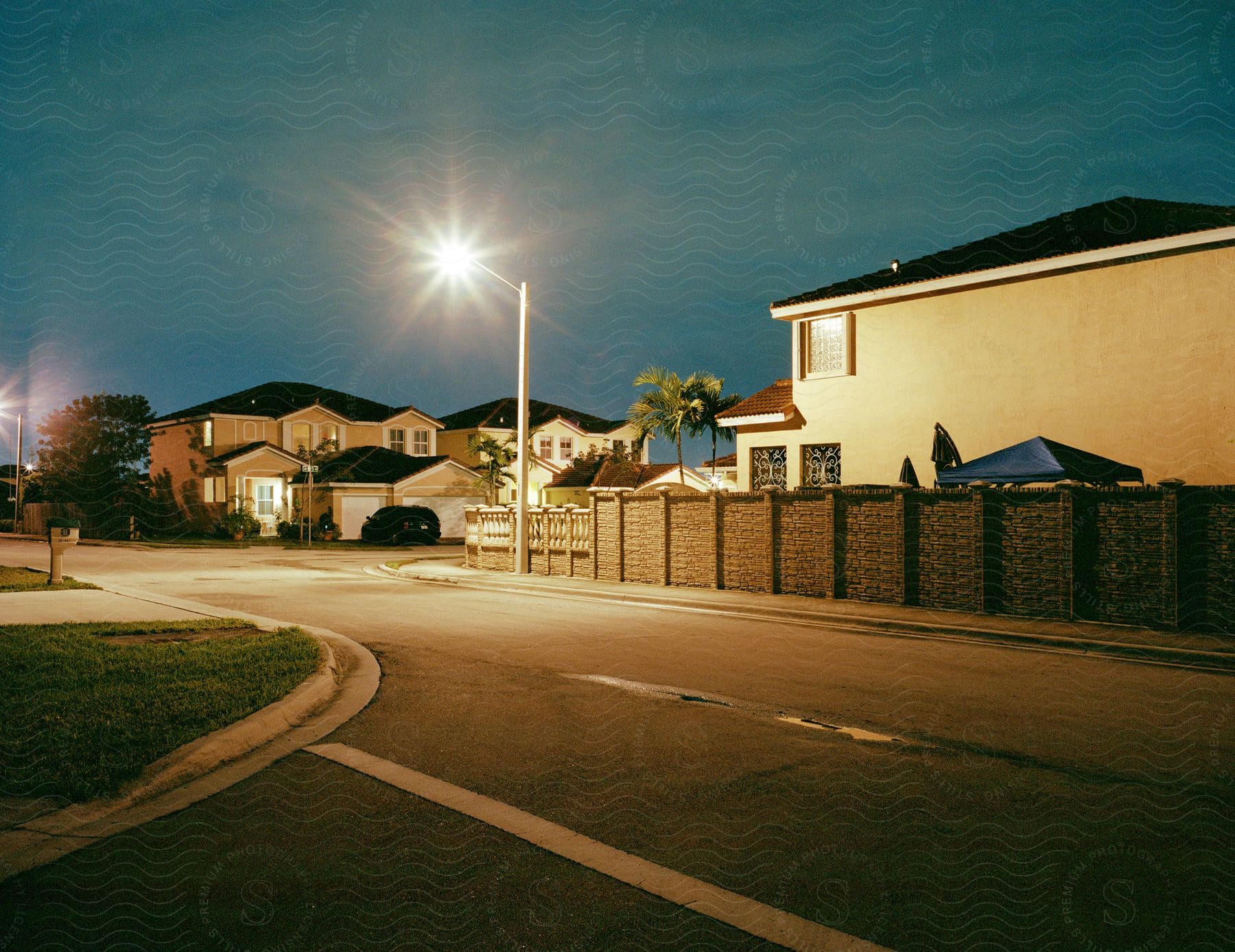 Curved road near fence with glowing street lights in a neighborhood with houses and trees