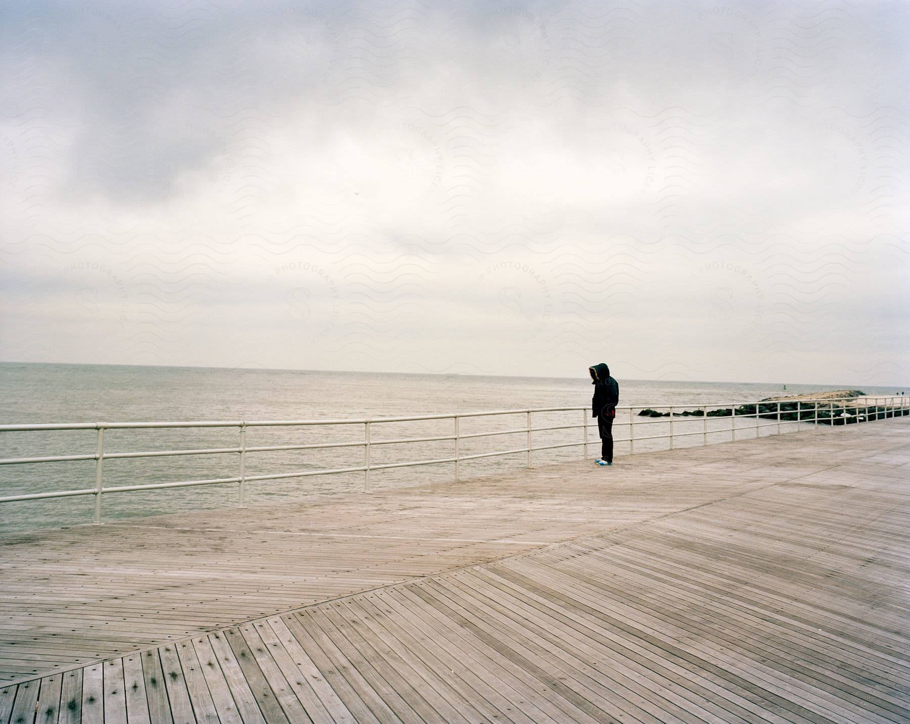 Man On Boardwalk Looking At Water Over Railing Wearing Jeans And Coat On Cloudy Day