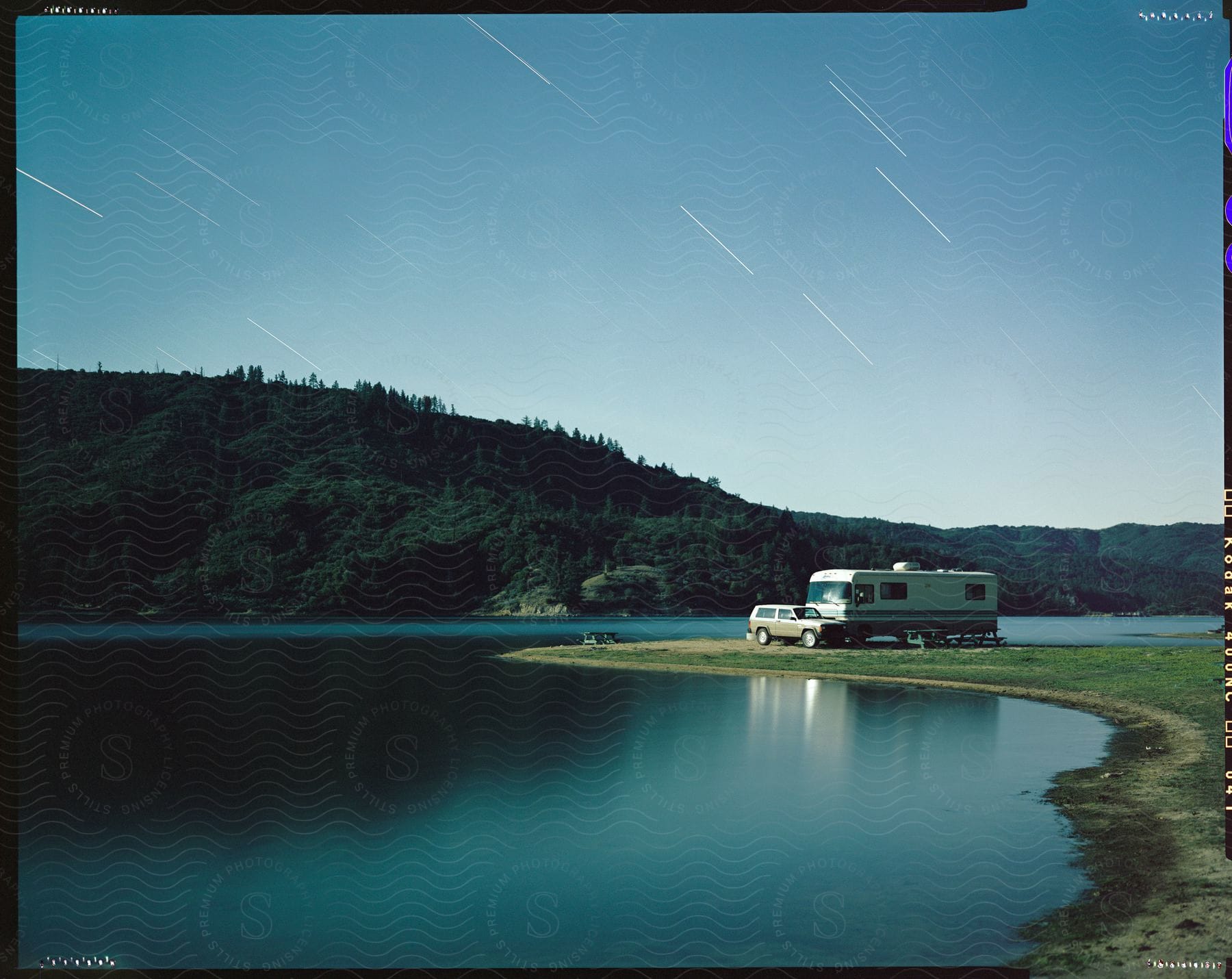 Meteor streaks hover over rv parked across water from forested mountain