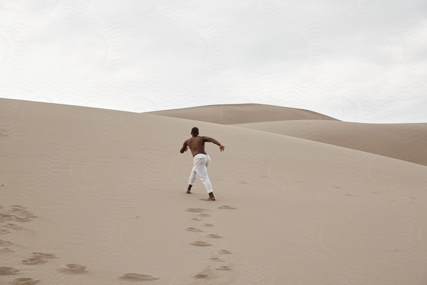 A shirtless black man wearing white trousers walking up a sand dune in the desert