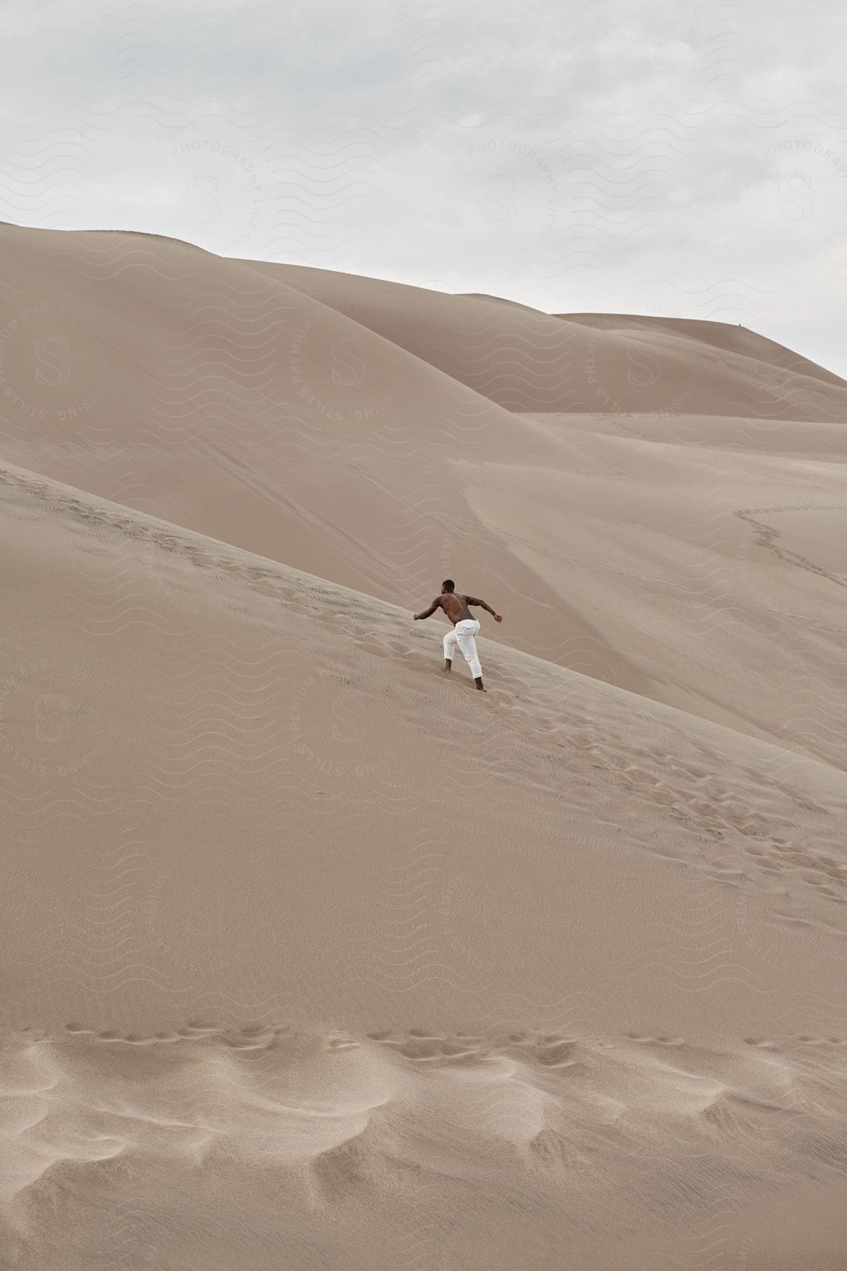 Shirtless man in white pants climbs sand dune in desert during the daytime