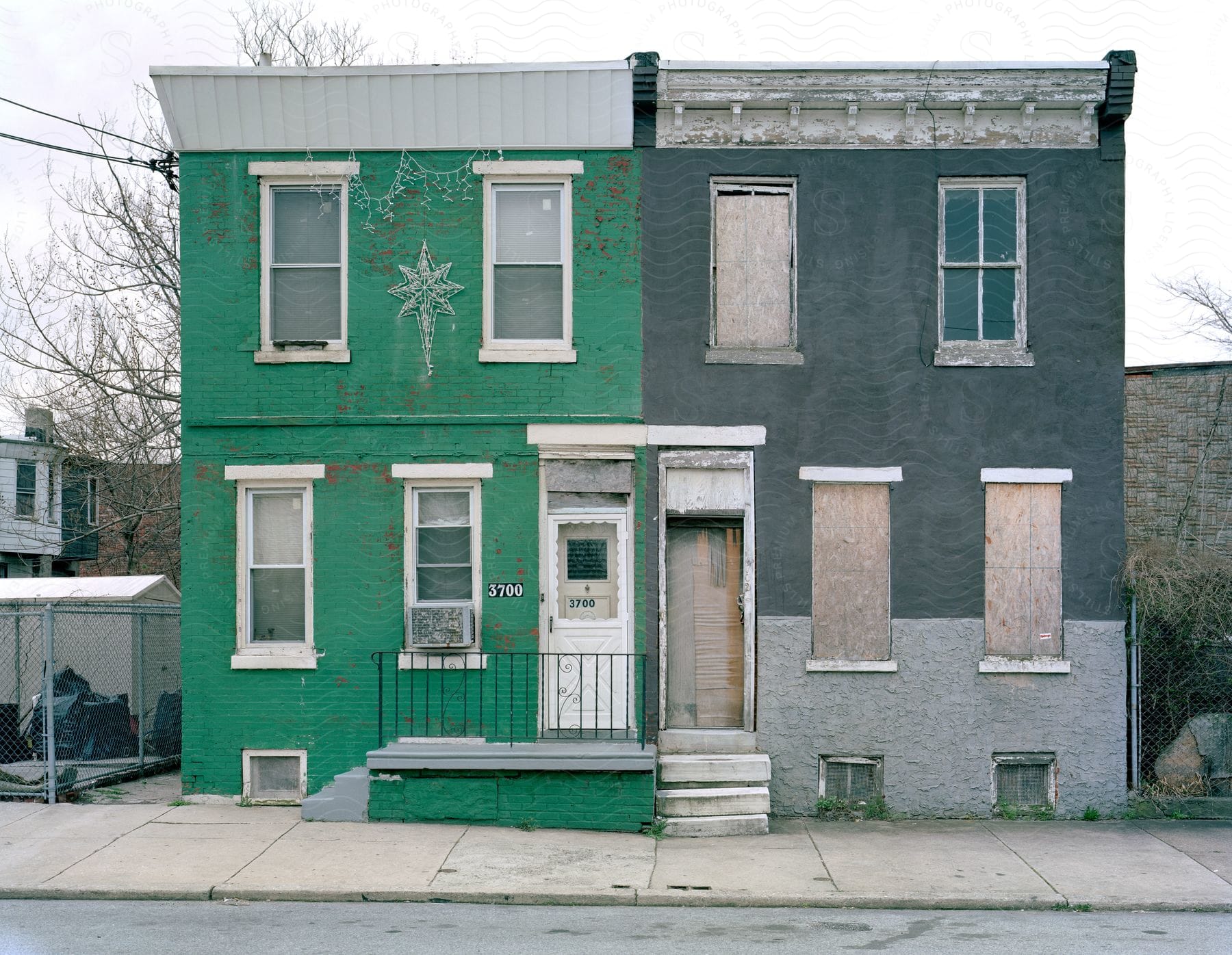 A green townhouse attached to a grey abandoned townhouse next to a sidewalk and a street in a residential area