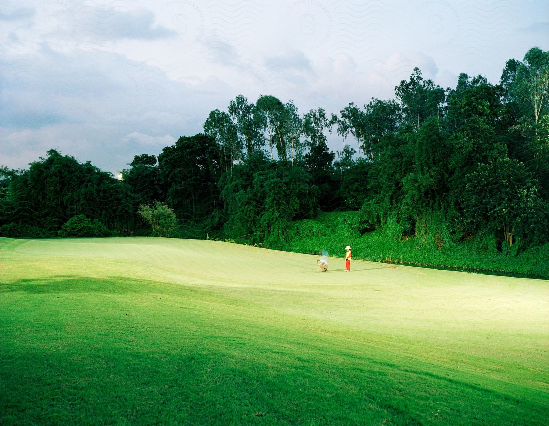 Stock photo of person stands while another person crouches on edge of golf course near woods