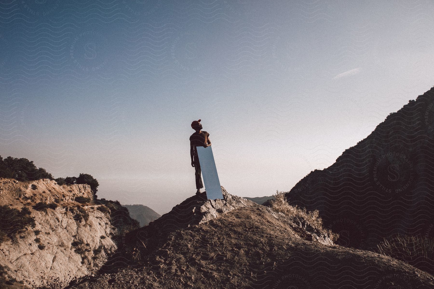 A person standing on top of a hill holds a mirror looking up at the sky