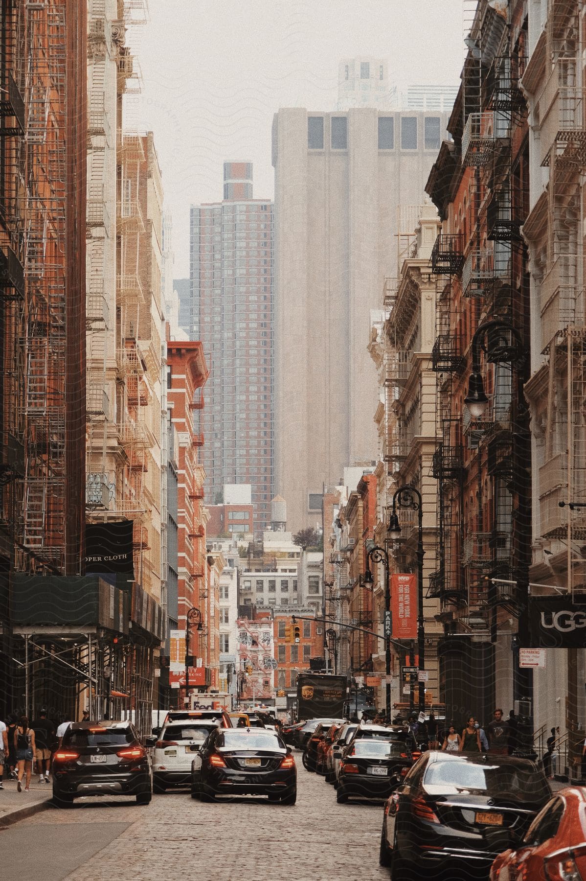 City street with cars parked and driving people walking on sidewalks in front of stores on a hazy day with tall buildings in the distance