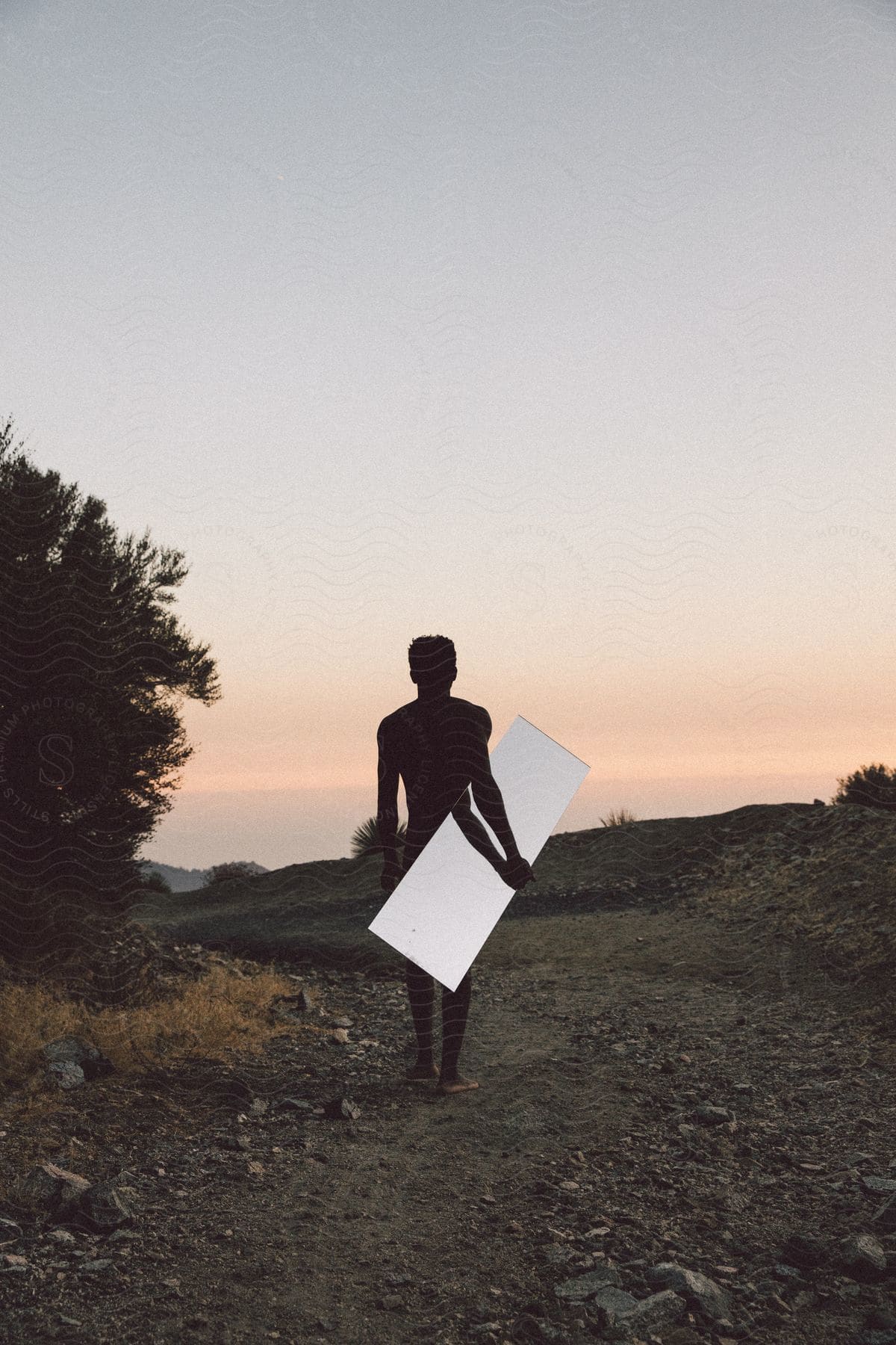 A barefoot man walks on a dirt road carrying a mirror in the hills at dusk