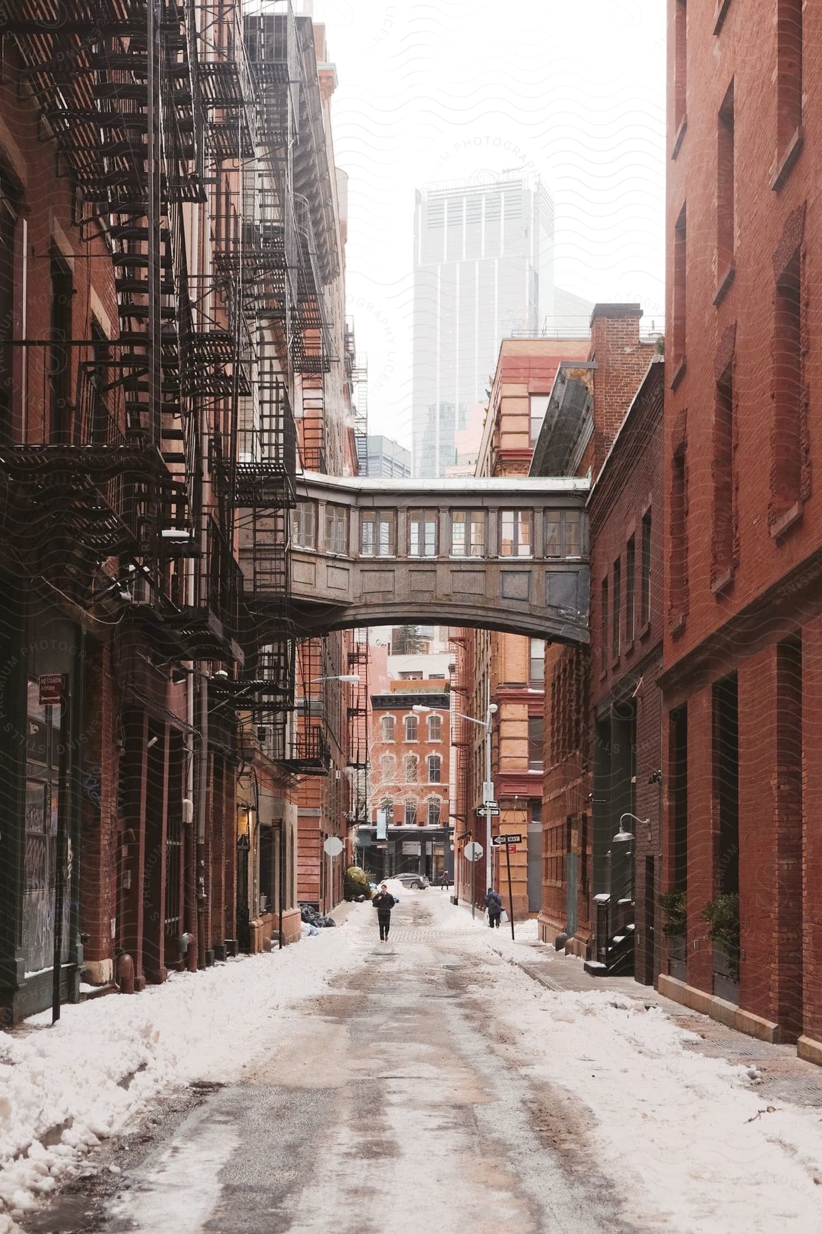 A snowcovered street with apartment buildings and a walk bridge connecting them a man stands still while another walks down the street