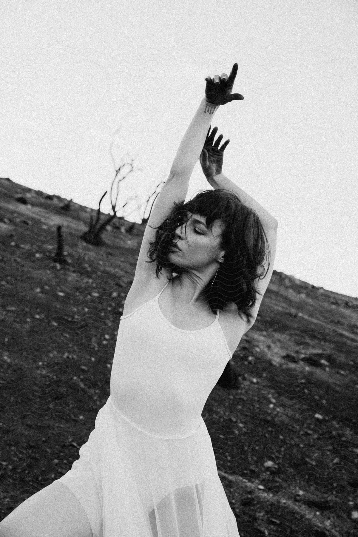 A woman in a white ballet dress stands in front of a barren wasteland with black paint on her palms and wrists raised arms towards the sky