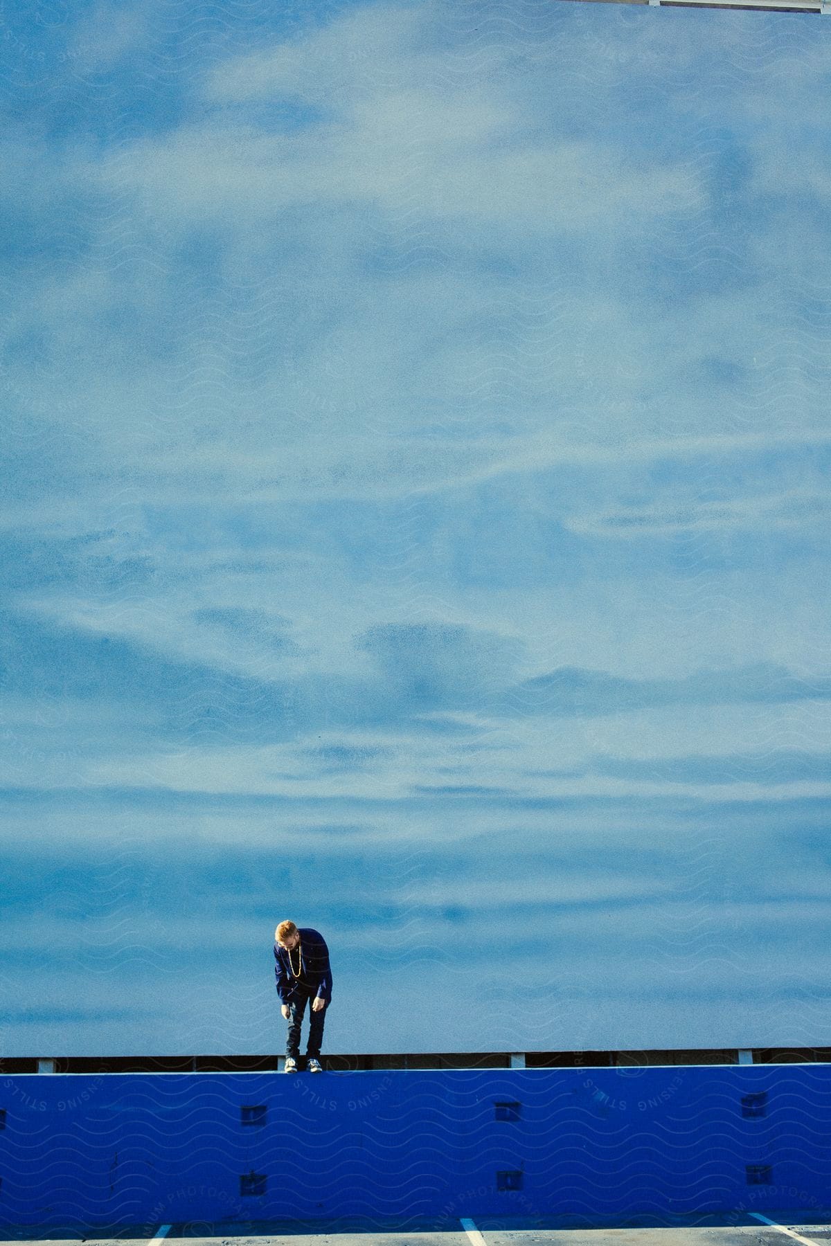 A man checking his shoe on a vibrant blue object on the street with a vast expanse of sky behind him