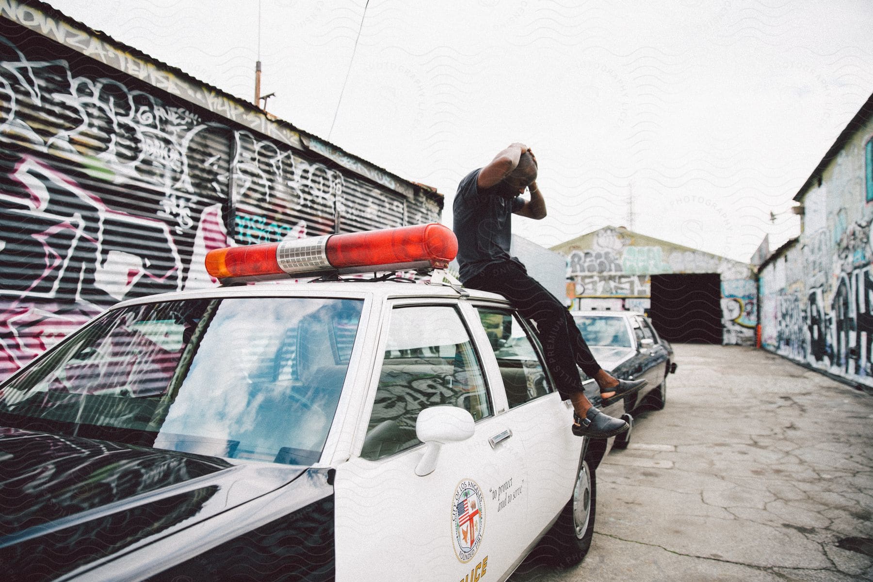 A man in a black tshirt pants and shoes sits on a police car with his hands behind his head