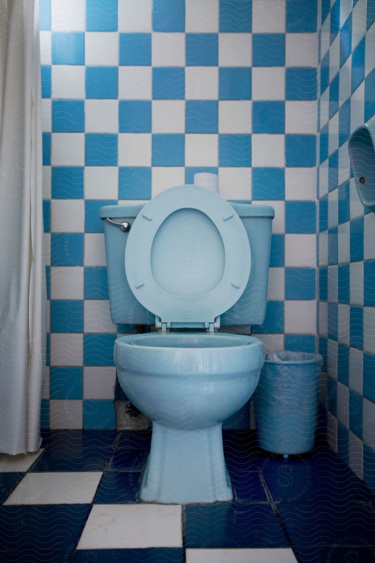 A light blue toilet with a wastebasket and blue and white tiles in a bathroom