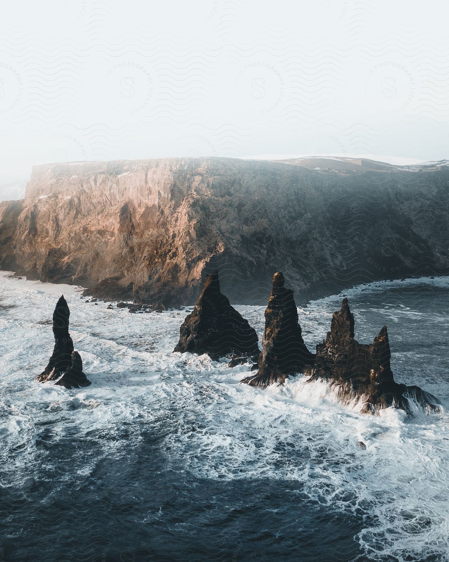 Sharp rugged rock formations jut out of the ocean amidst whitewater and mist framed by a rock cliff