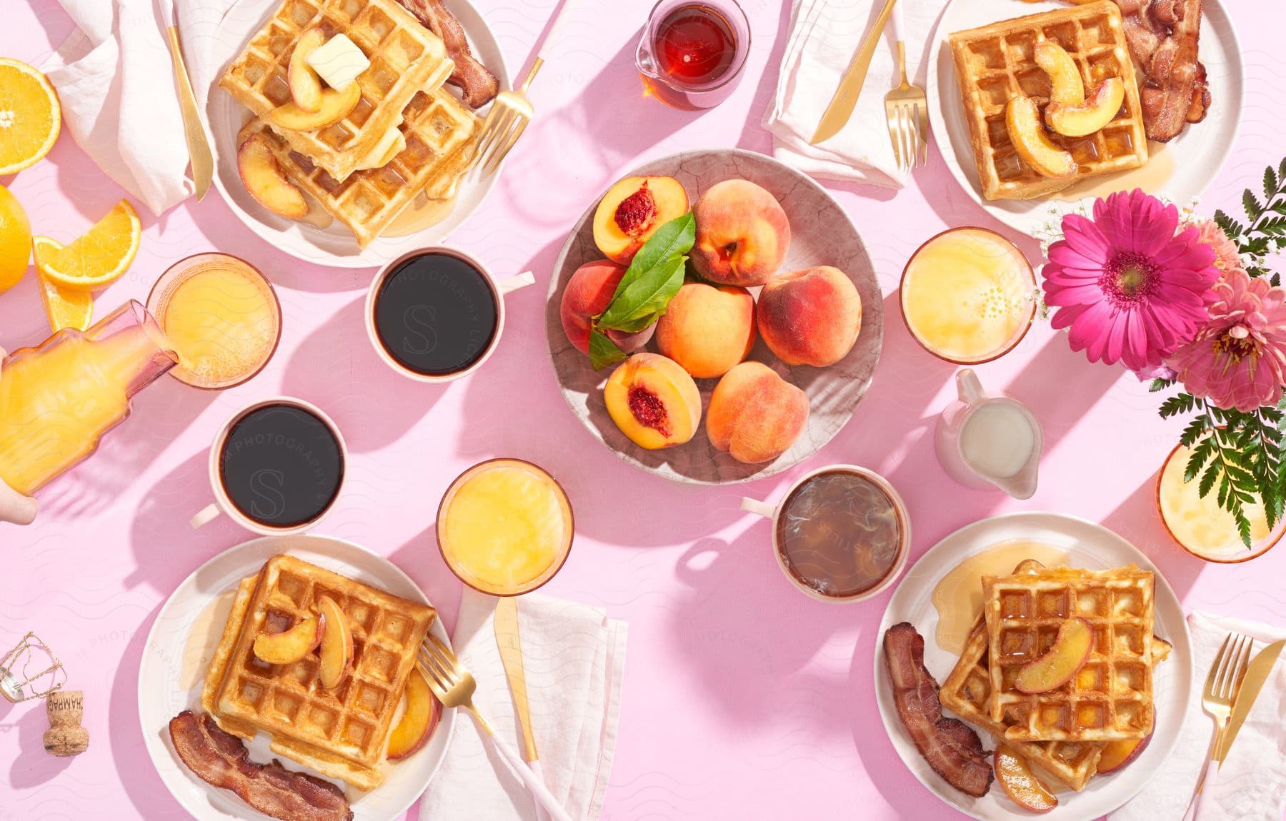 Person pouring orange juice into a glass on a pink table with various food and drink items