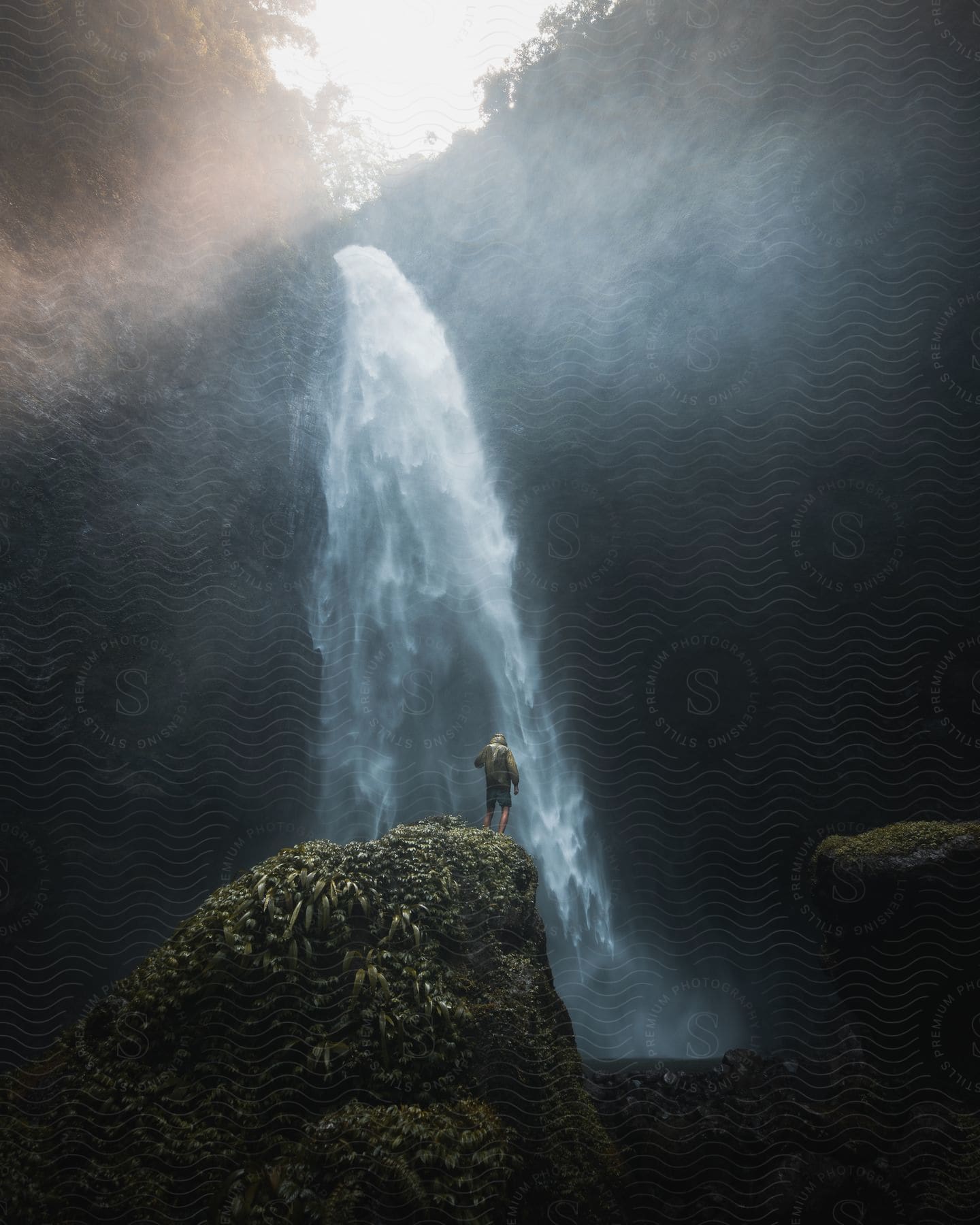 A person stands on a mountain facing a majestic waterfall amidst beautiful scenery