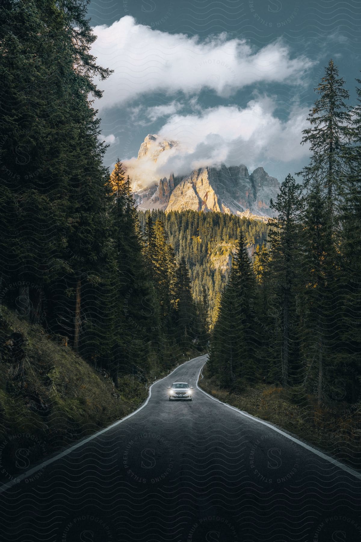 A white car with headlights on drives on a forest road with mountains in the distance under a cloudy sky