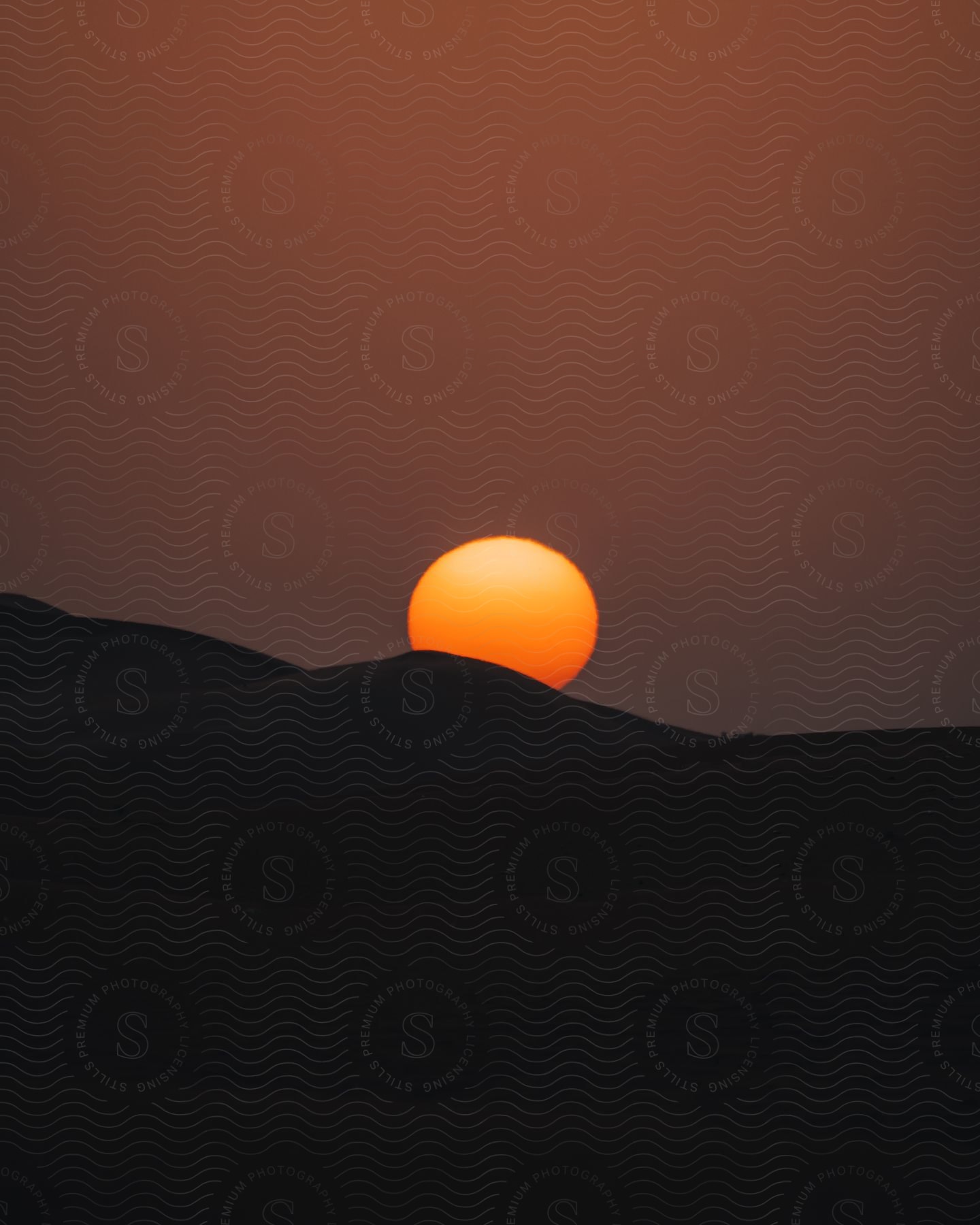 A glowing sun sets over a mountain in a deep dark red sky during dusk