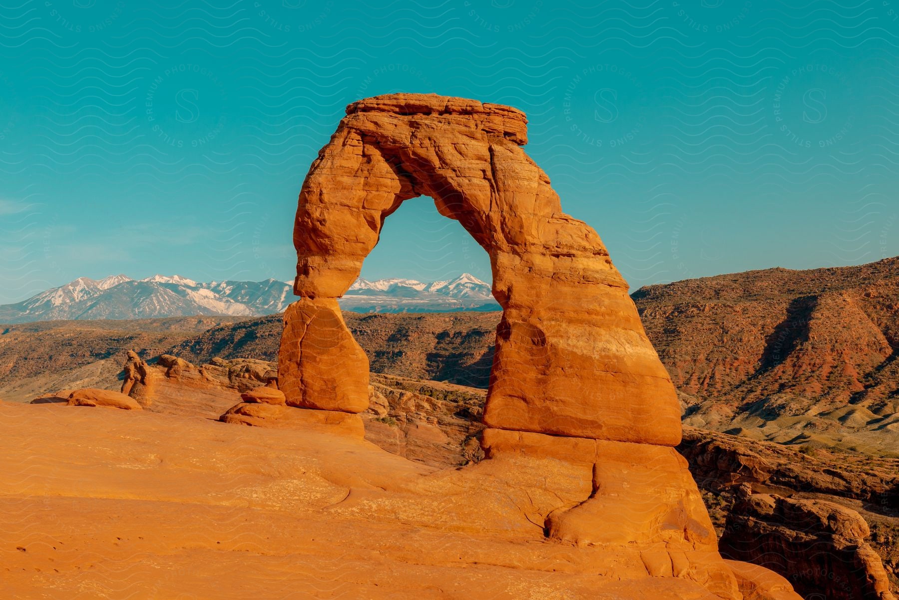 Stock photo of red rock arch shines in desert badlands with mountains in the distance under a clear blue sky