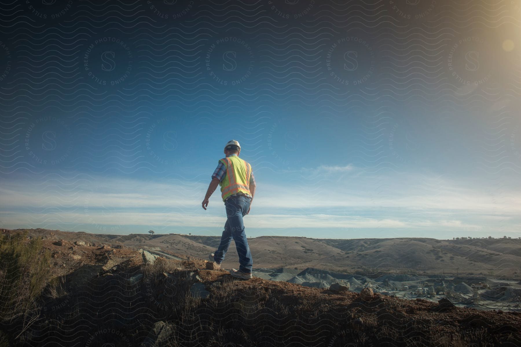 Worker walking across ridge overlooking pit wearing vest and hard hat