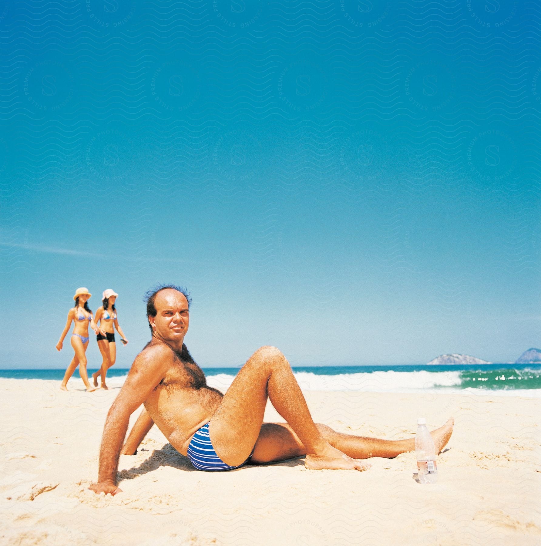 Two females in bikinis walking and one man in swim trunks sitting on the beach under a clear blue sky