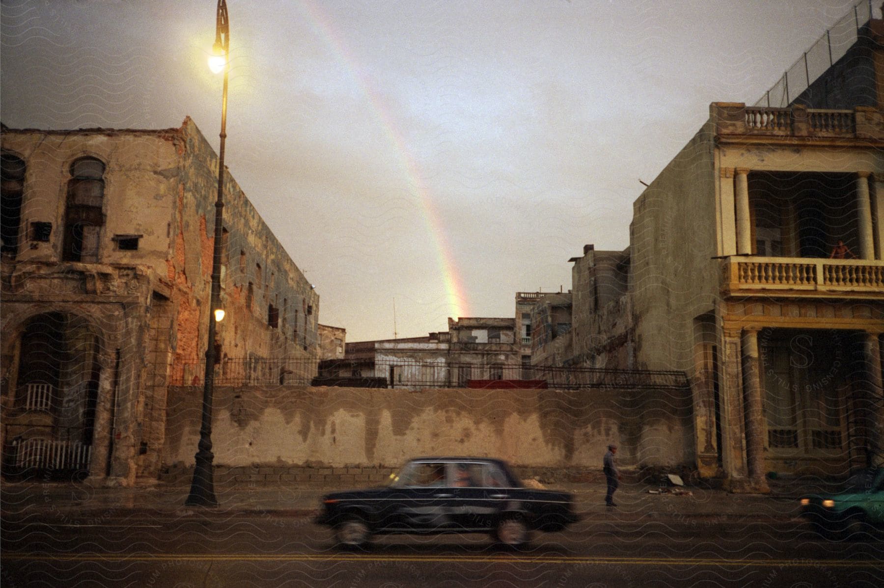 A black car drives down a street passing old buildings as a rainbow appears in the background