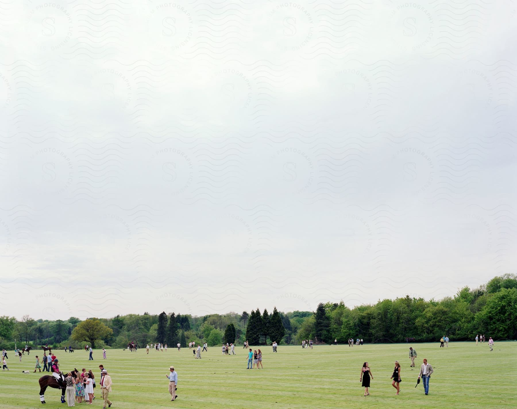 A crowd of people and polo players on horses walk on a polo field on a cloudy day