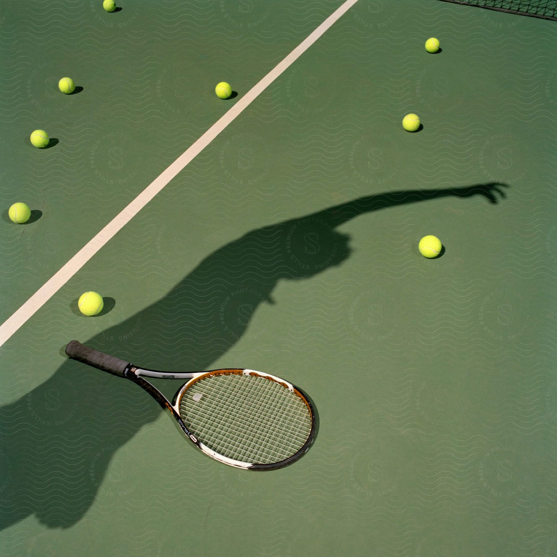 A tennis court with scattered balls a rack and the shadow of a player