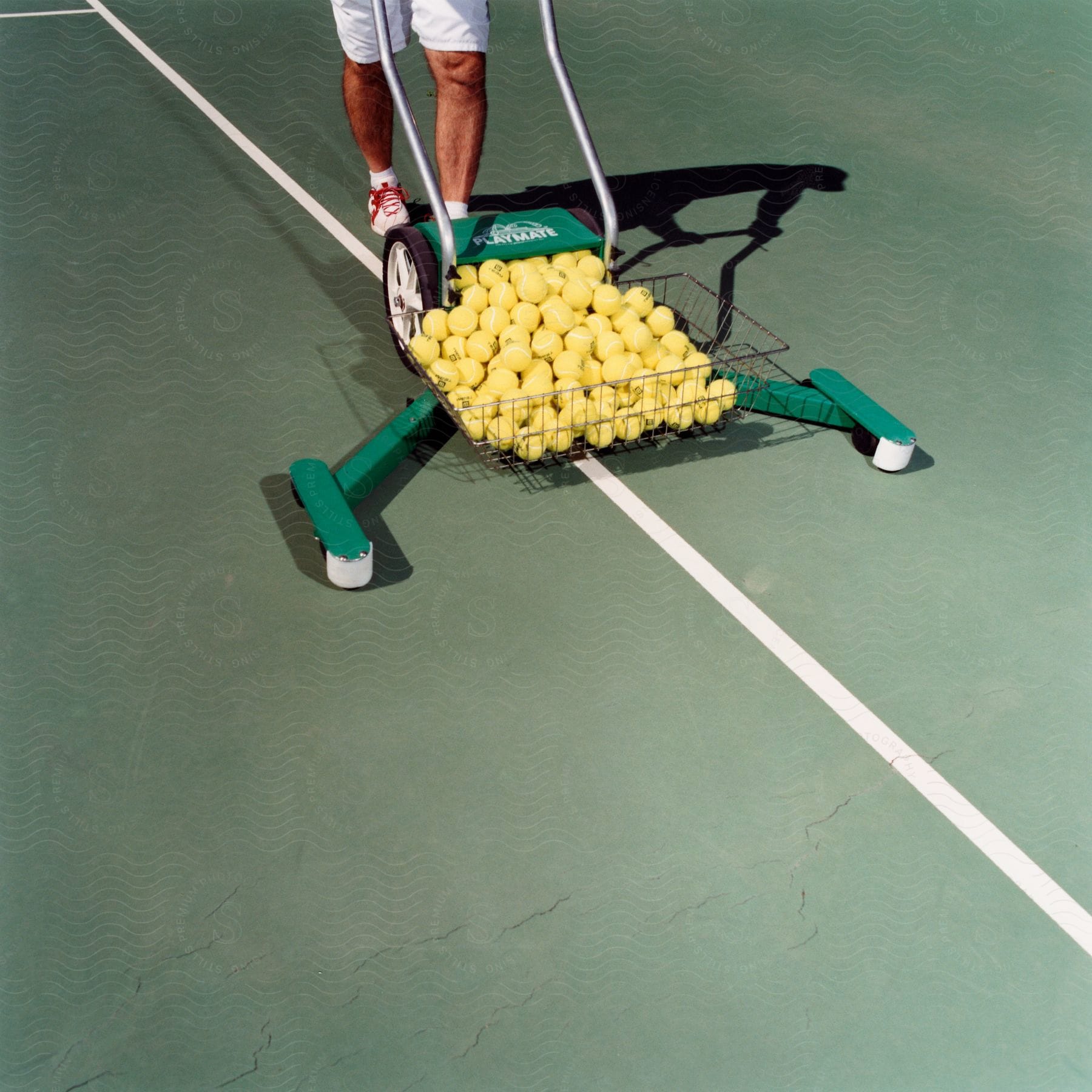 A Person In White Shorts Is Using A Tennis Ball Mower To Collect The Balls From The Court