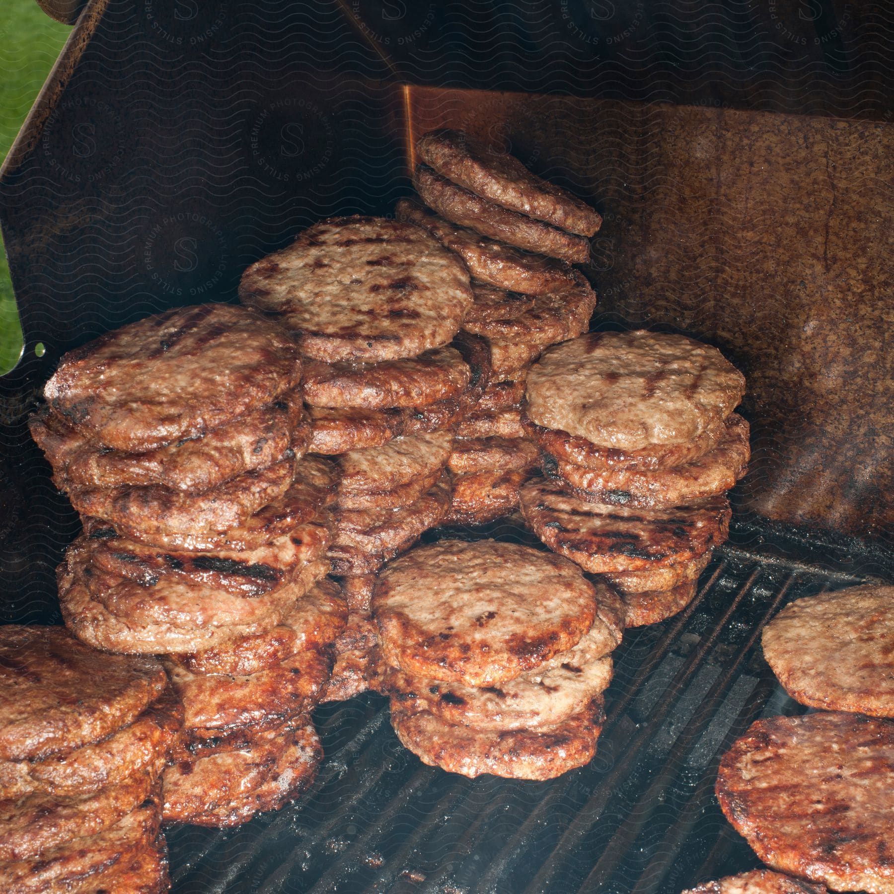 Dozens of cooked hamburgers are stacked on a grill outside