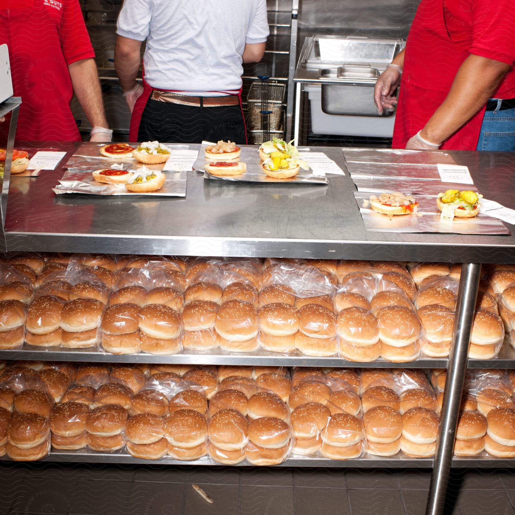 People preparing hamburgers at a metal counter with various buns on a shelf