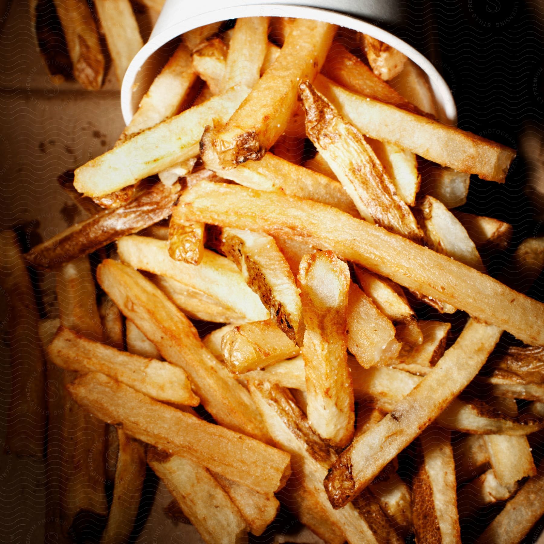 A cascade of golden brown fries spills out of a paper cup and into a brown paper bag
