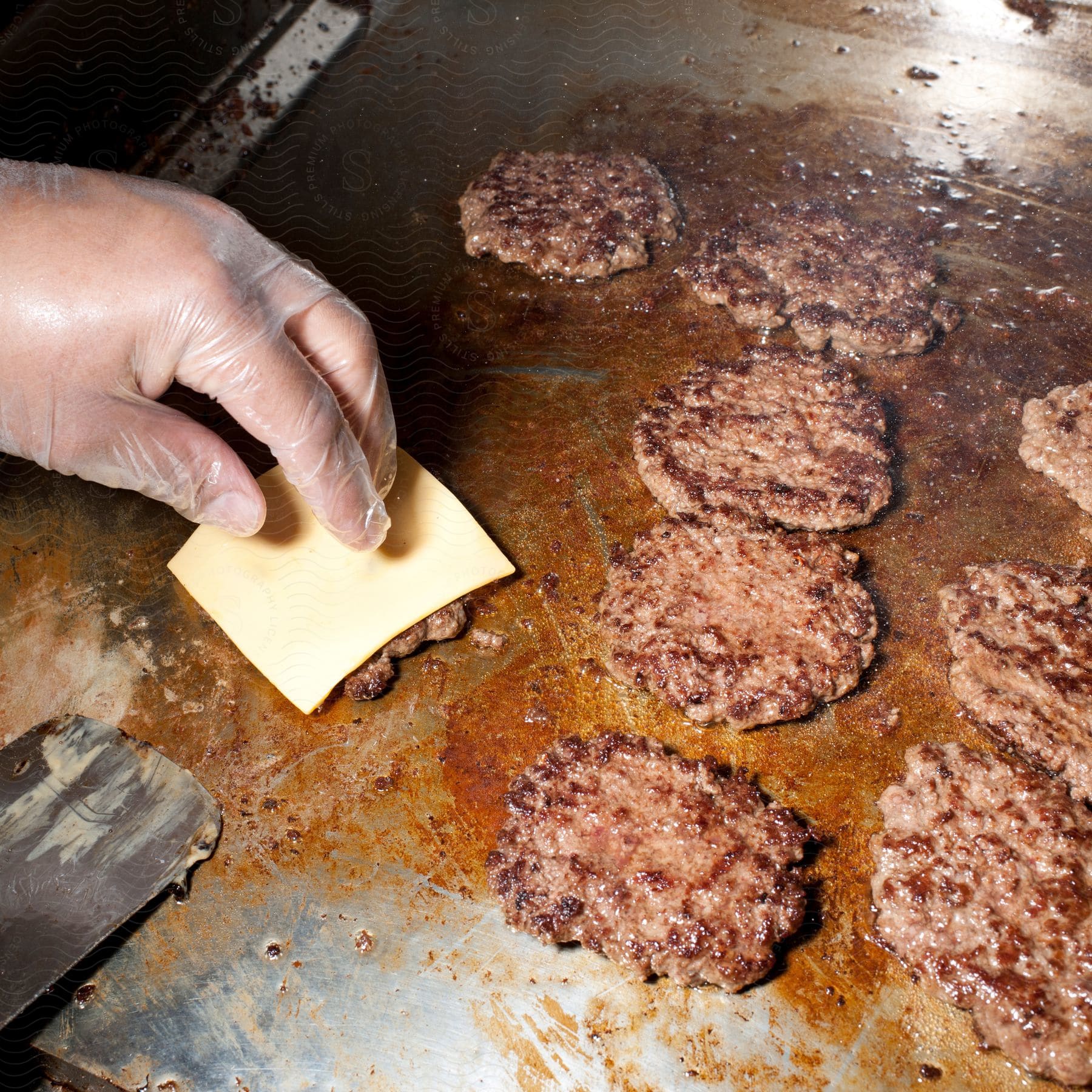 A chef adds a slice of cheese to a cooking burger on a commercial stove surrounded by other burgers
