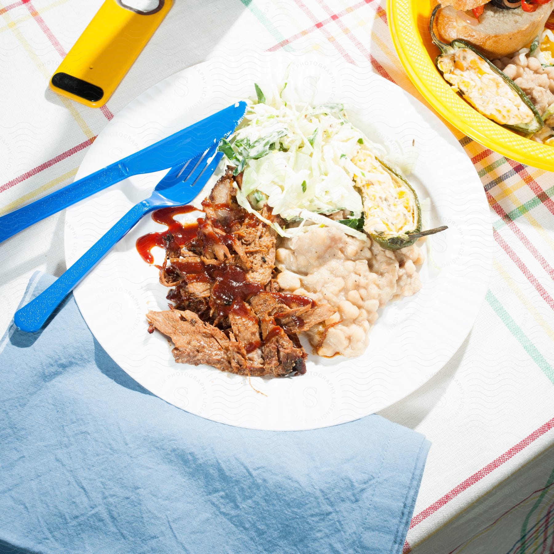 Barbecue food on a paper plate with blue utensils on a colorful tablecloth with a blue napkin