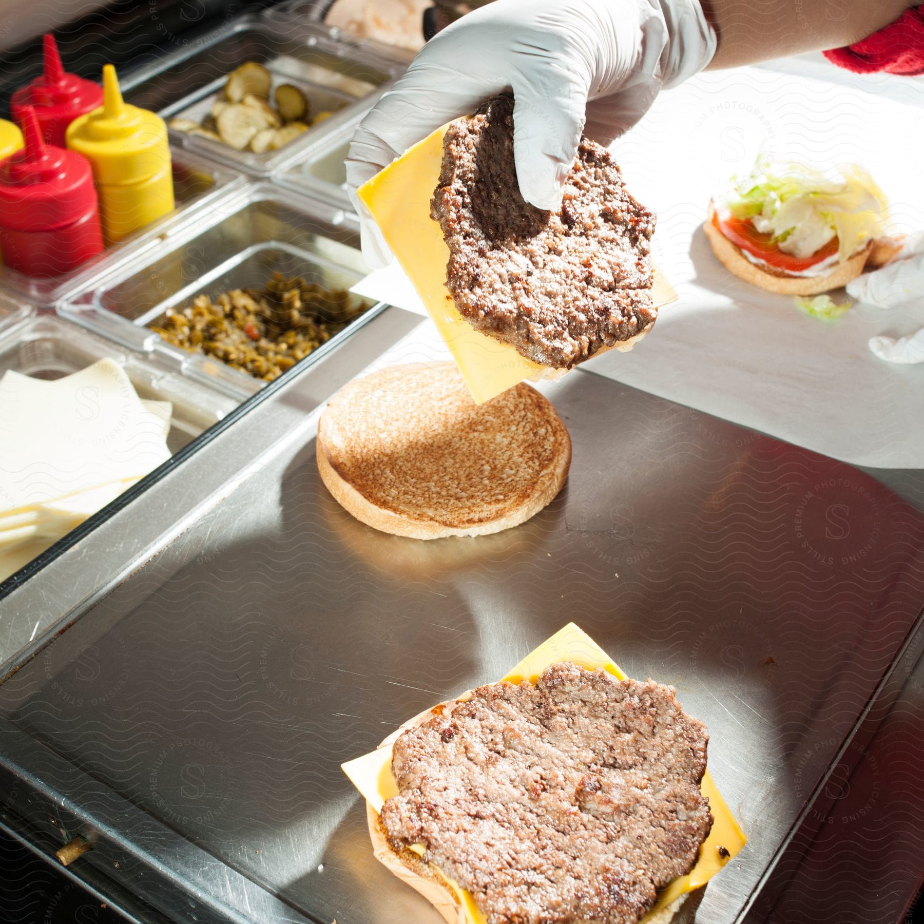 Hand placing a meat patty with cheese over a bun with other ingredients like lettuce pickles and sauces
