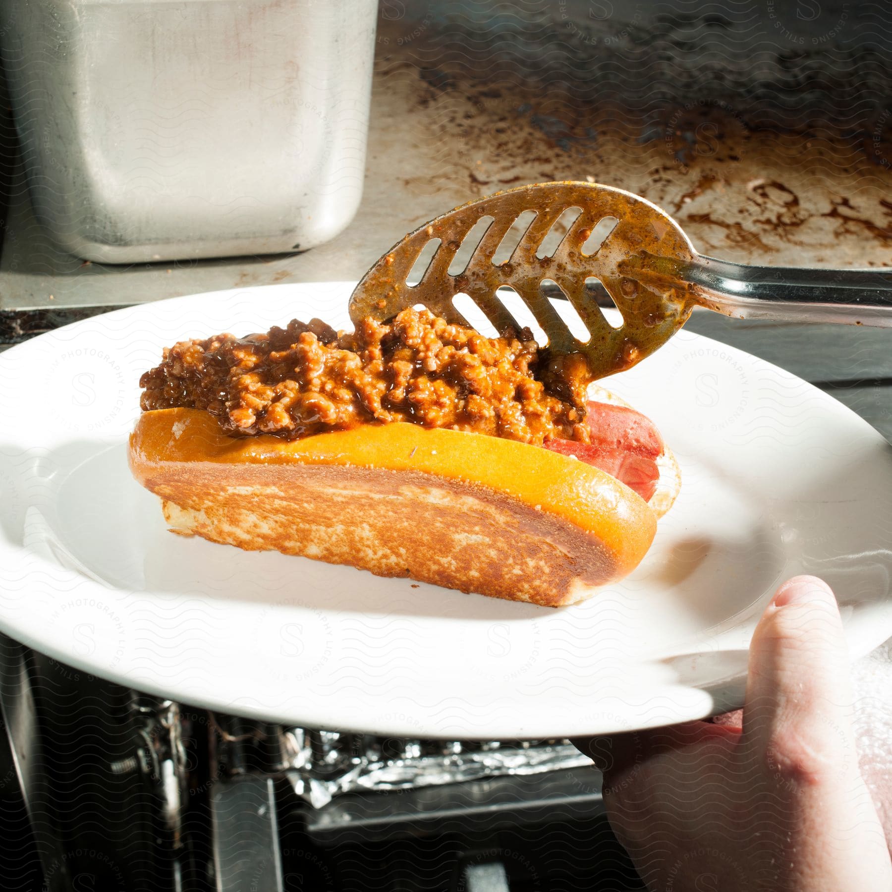 A person garnishes a chili dog with a generous helping of chili in a kitchen