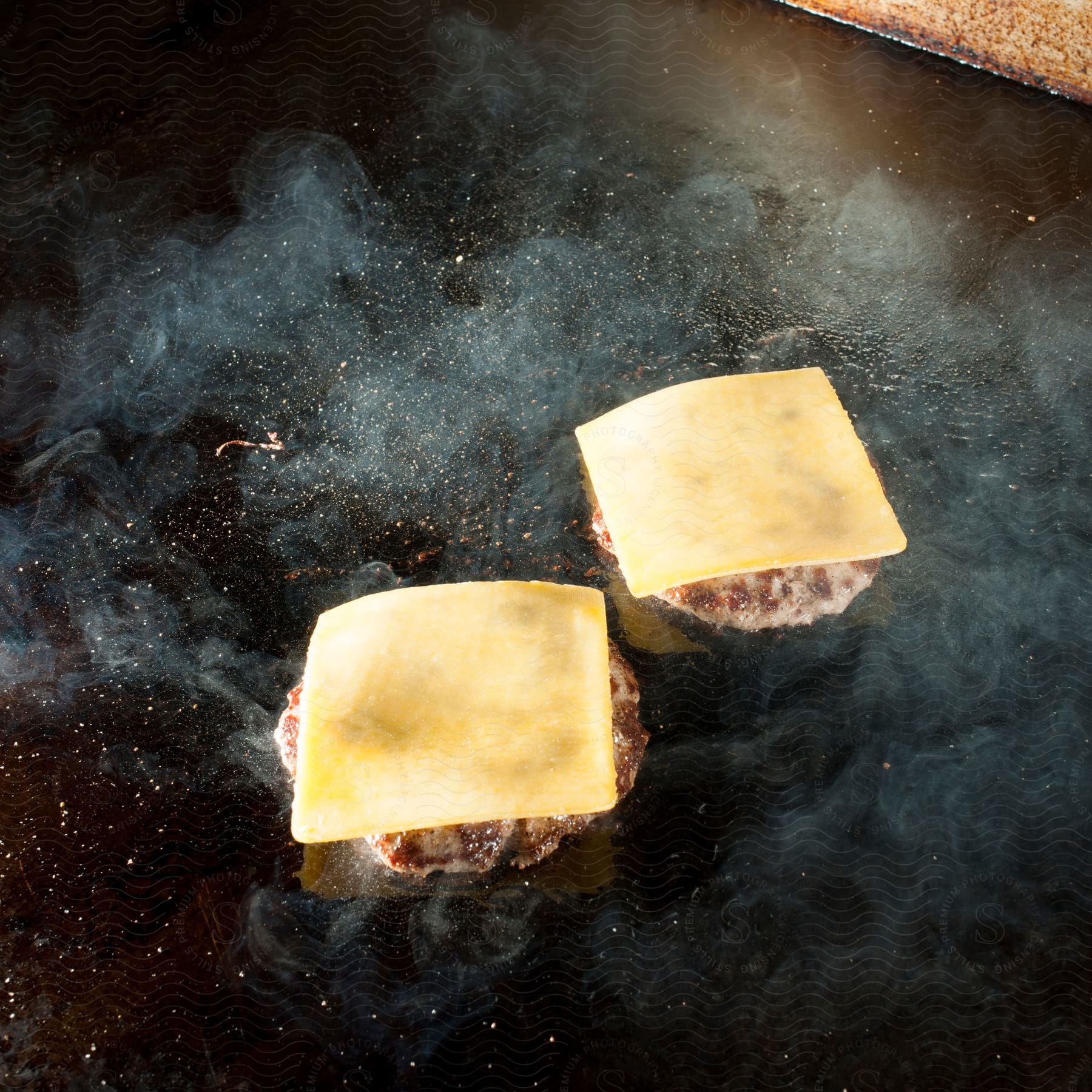 Two burgers cook on a grill with melting cheese on top