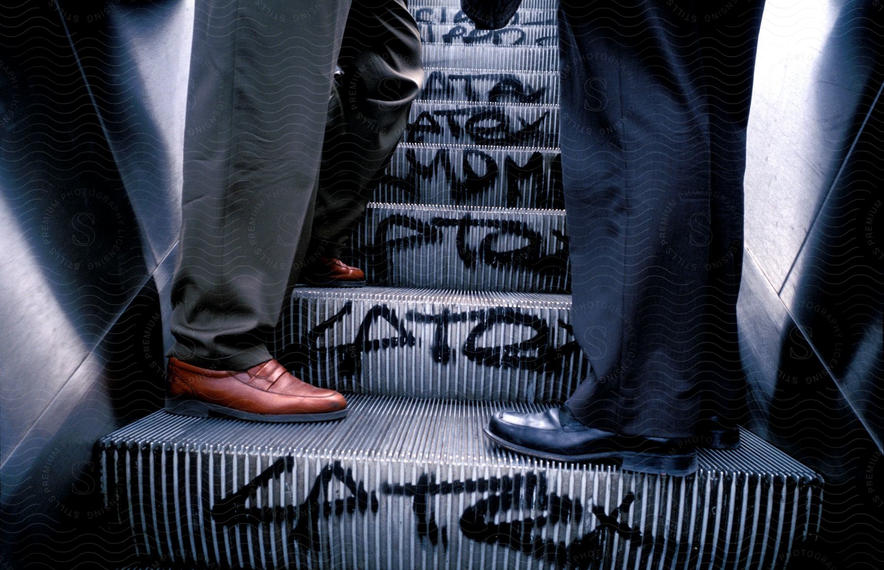 Two people wearing formal pants and shoes standing in front of each other on an escalator with graffiti