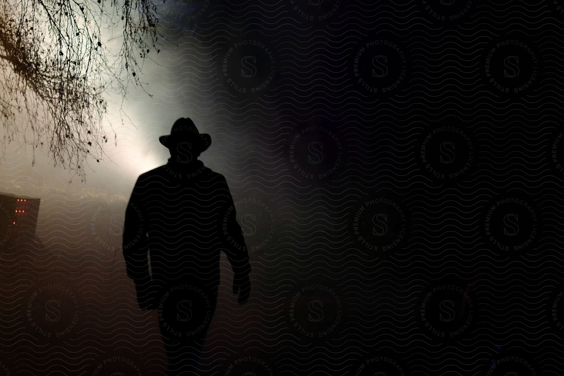 Stock photo of a man in a hat walks through a foggy field silhouetted by a light behind him with farm equipment and a leafless tree in the distance