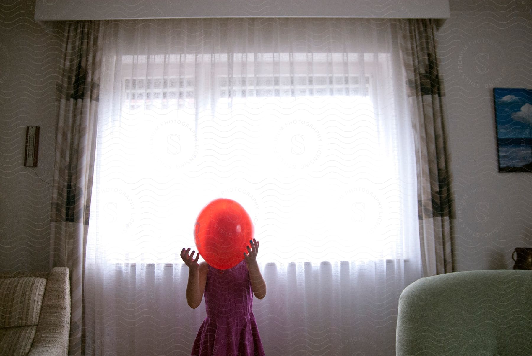 A young girl in a purple dress holds a red balloon in front of her face illuminated by sunlight streaming through living room curtains