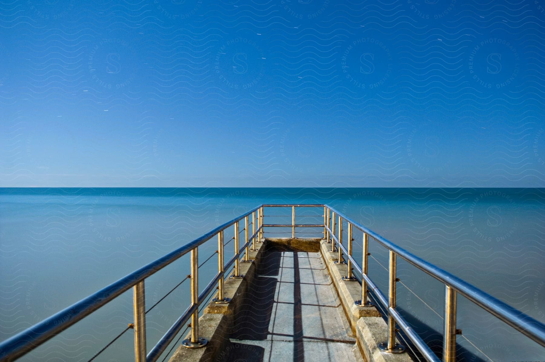 A sunlit pier with a metal railing under a blue sky