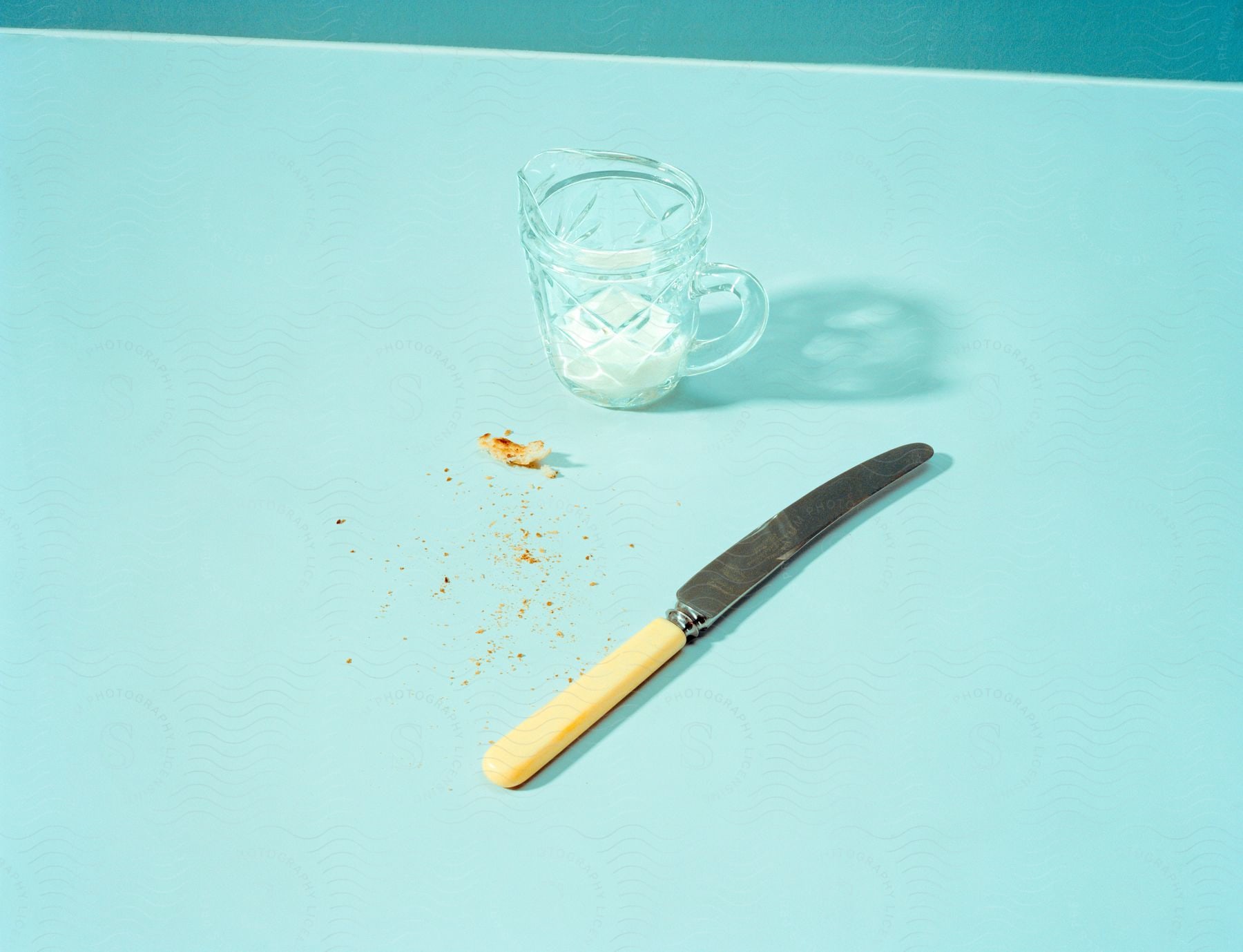 Butter knife with yellow handle laying by crumbs and glass cup on teal surface