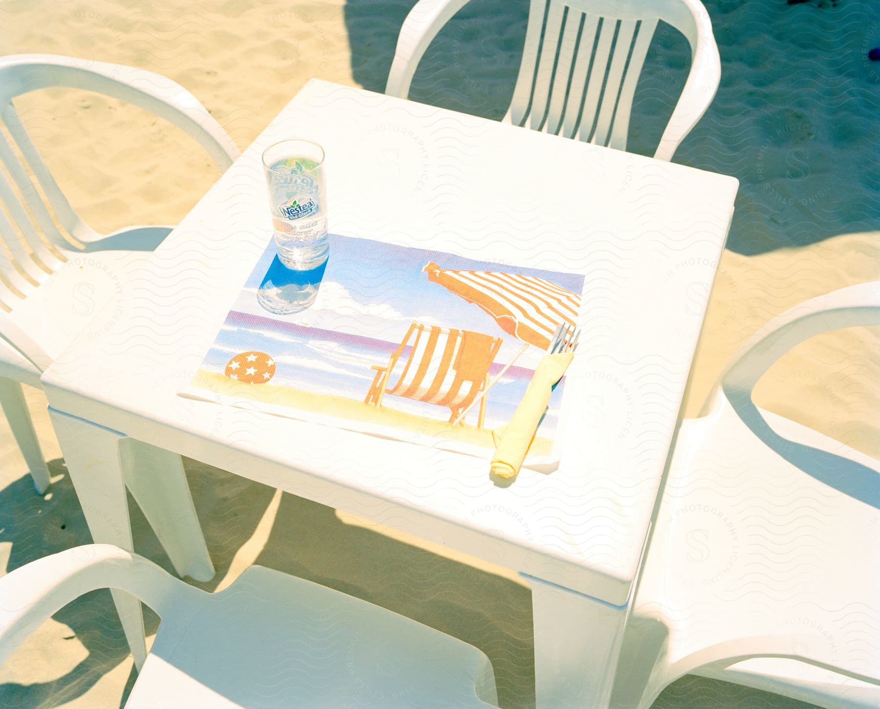 Four white plastic chairs around a square white plastic table with a beach lawn chair scene placemat and a glass of water on the beach beside a beach umbrellas shadow on a bright summer day