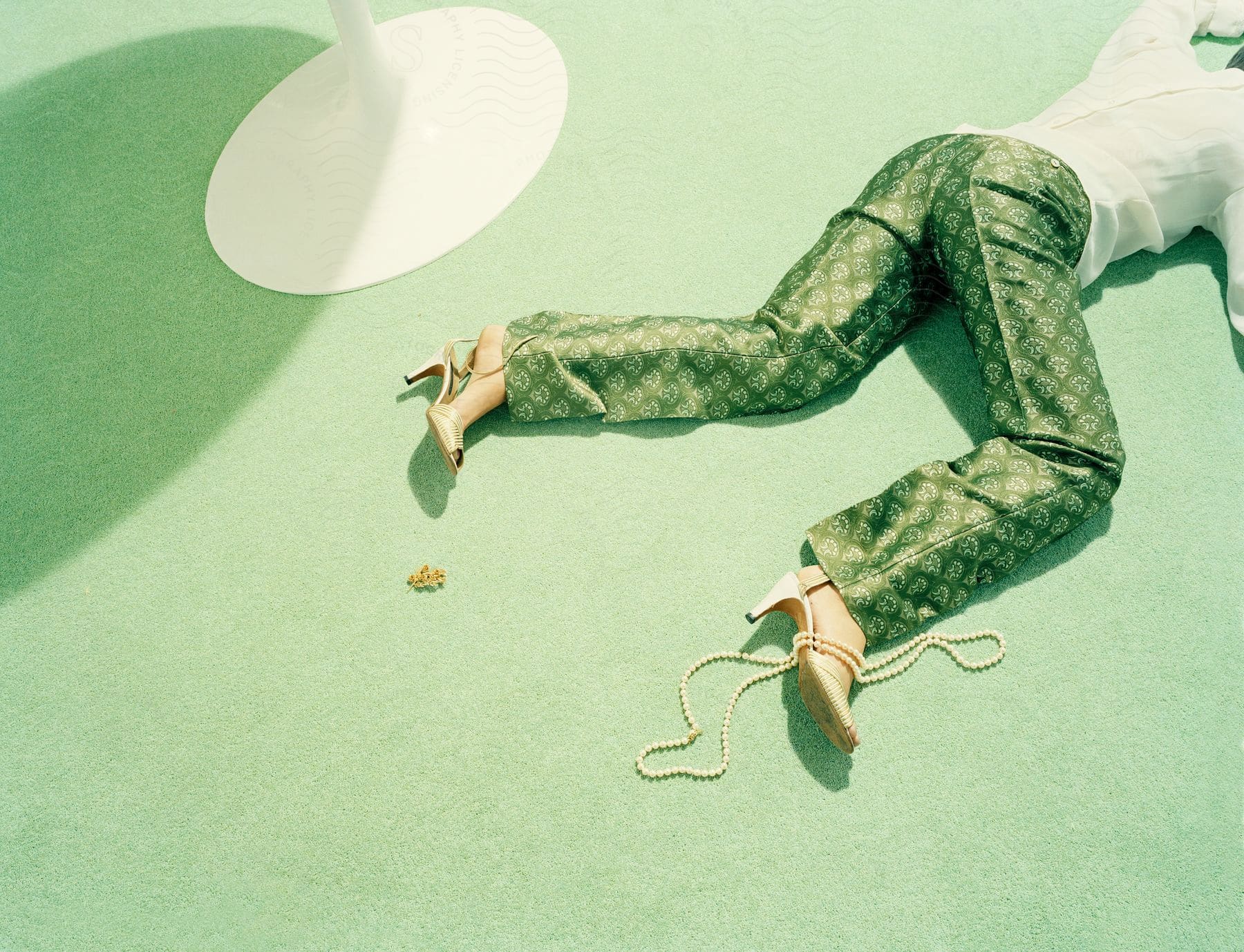 A woman in green pants and a white shirt lying on the floor next to a white table with a string of pearls over her right foot