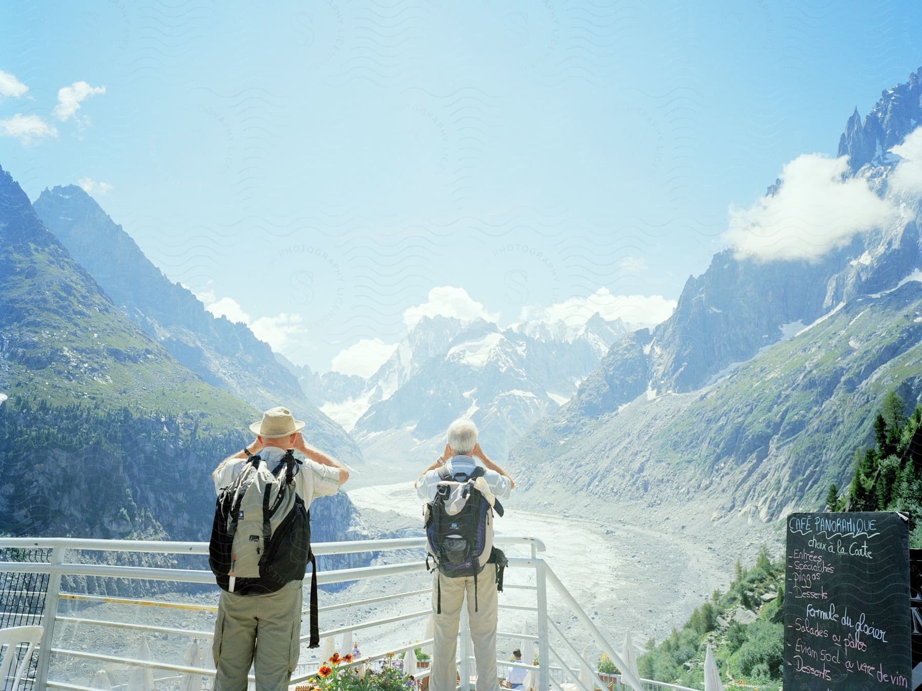 Two hikers stand on a walkway overlooking mountains and a river valley taking photographs