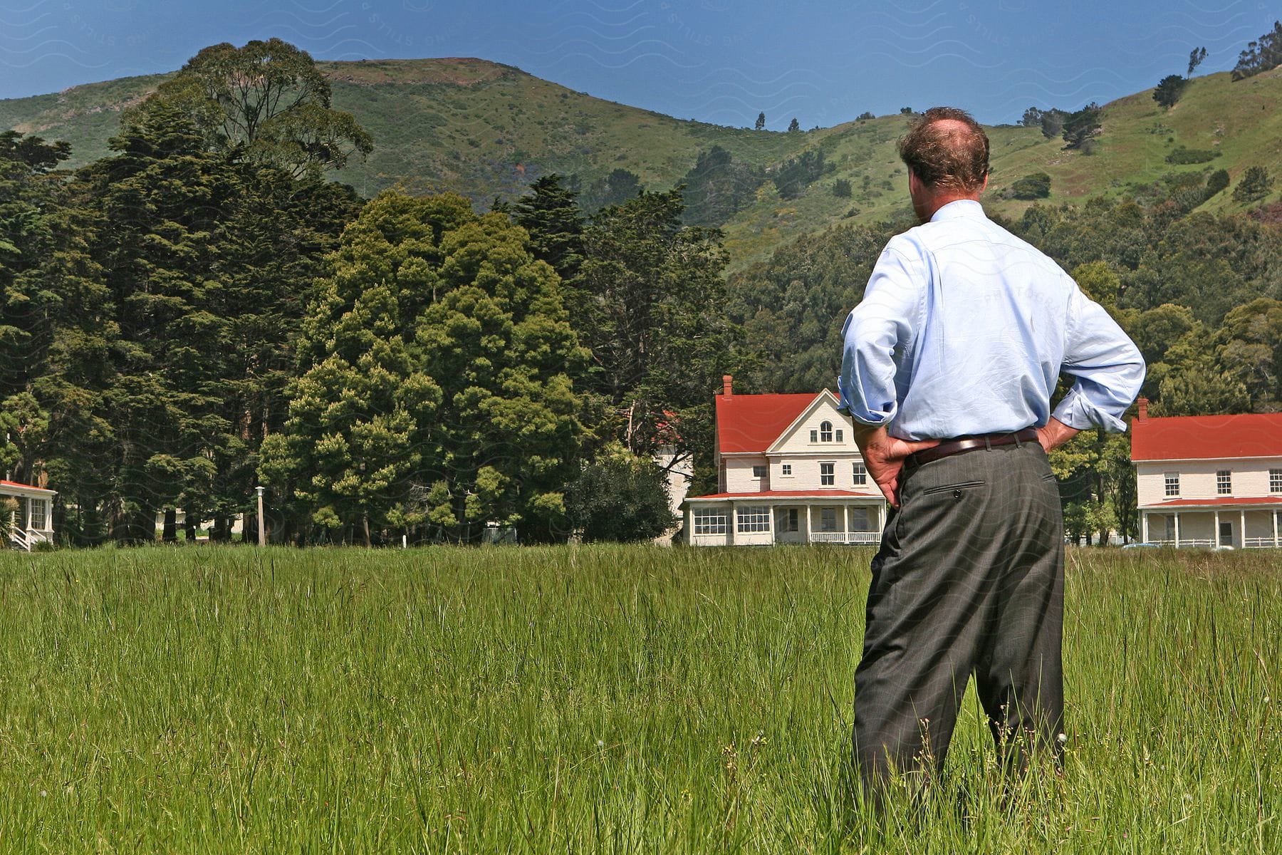 A man stands in a field looking at a house in the distance with mountains and trees in the background