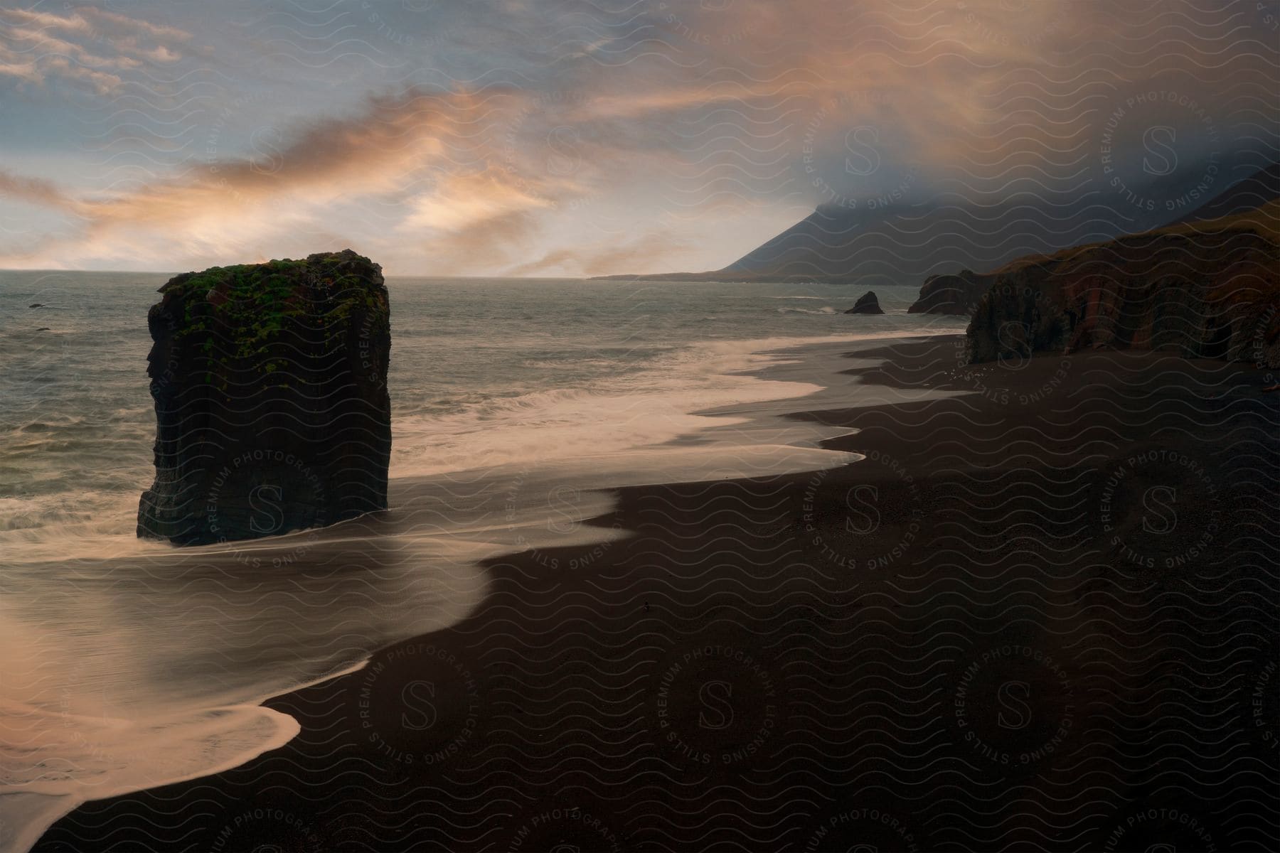 A square shaped rock stands in the ocean on a sand beach at dawn